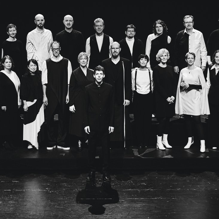 Das Vokalensemble steht positioniert für ein Gruppenbild auf einem Holzboden. Ein Mann steht vor den Reihen der anderen. Das Bild ist schwarz-weiß.