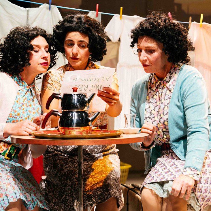 Drei Frauen sitzen auf der Bühne an einem kleinen Tisch, lesen gemeinsam ein Dokument, Wäsche im Hintergrund