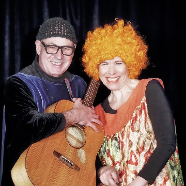 Christof und Vladi Altmann auf der Bühne. Er trägt ein langes lilafarbenes Kostüm und hat eine Gitarre in der Hand. Sie trägt ein buntes Kleid und eine Perücke mit kurzen orangefarbenen Locken.