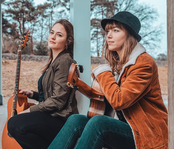 Portaitfoto Sophie Chassée und Karlijn Langendijk. Sie sitzen draußen unter einem Vordach und haben beide Gitarren in der Hand.