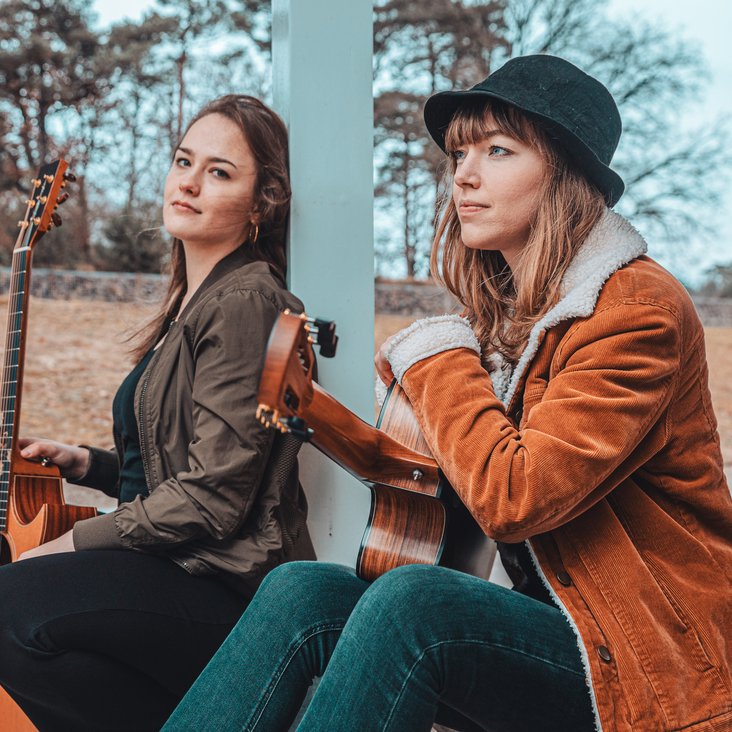 Portaitfoto Sophie Chassée und Karlijn Langendijk. Sie sitzen draußen unter einem Vordach und haben beide Gitarren in der Hand.