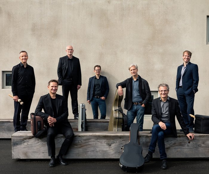 Gruppenporträt eines Ensembles vor einer steinernen Wand, Musikerinnen und Musiker in formeller Kleidung.