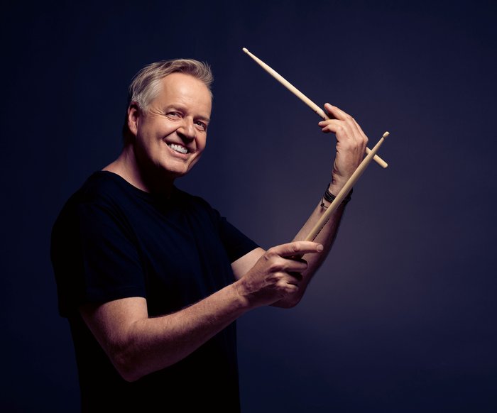 Portraitfoto Wolfgang Haffner, er trägt ein schwarzes T-Shirt, blickt lächelnd in die Kamera und hat Drum Sticks in den Händen.