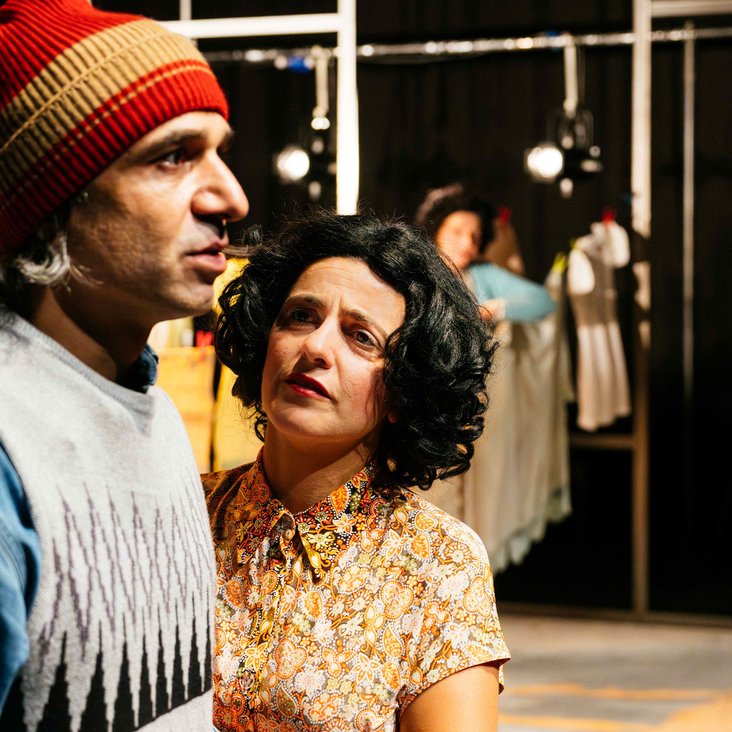 Frau und Mann stehen nah beieinander, Frau schaut skeptisch oder traurig-angestrengt, Bühnenbild mit Kleidung im Hintergrund