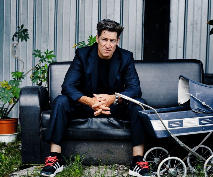 Tobias Moretti sitzt im Freien auf einer breiten Lederbank, neben ihm stehen Pflanzentöpfe und ein altmodischer Kinderwagen.