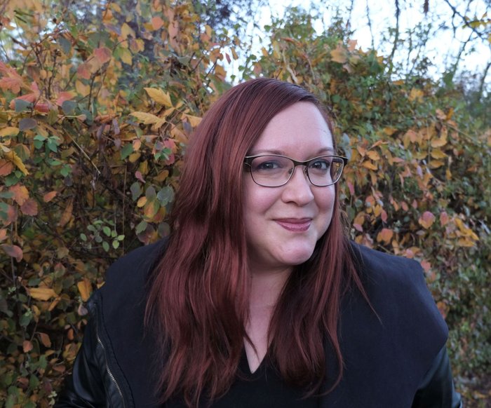 Porträt einer Frau mit rotem Haar und Brille im Freien, herbstliche Bäume im Hintergrund.