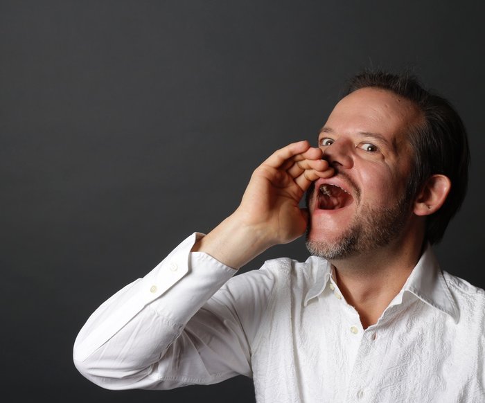 Porträt eines Mannes im weißen Hemd, der mit offener Hand ruft oder singt vor dunklem Hintergrund.