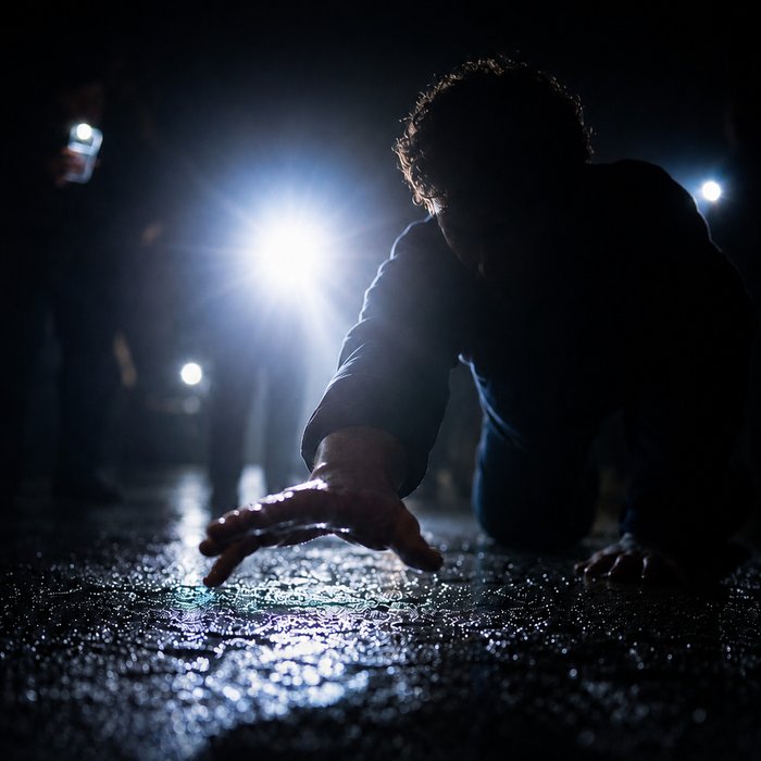 Kniende Person im Vordergrund streckt Hand über nassen Boden, grelle Lichter im Hintergrund.