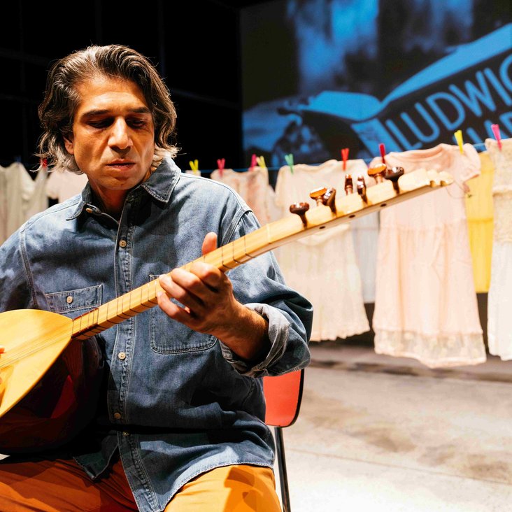 Mann spielt Saz vor einer Wäscheleine mit aufgehängten Kleidern auf der Bühne