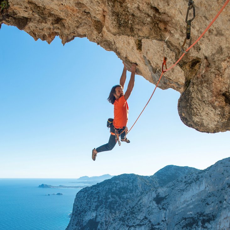 Alexander Huber hält sich mit beiden Händen an einem großen Felsvorsprung fest, im Hintergrund Felsküste und das Meer. Es ist mit einem Seil gesichert.