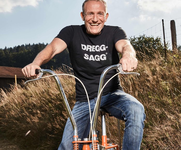 Michl Müller sitzt auf einem orangefarbenen Fahrrad, er fährt auf einer Wiese oder einem Feld Richtung Fotograf:in. Er trägt Jeans und ein schwarzes T-Shirt mit der Aufschrift "Dregg-Sagg"