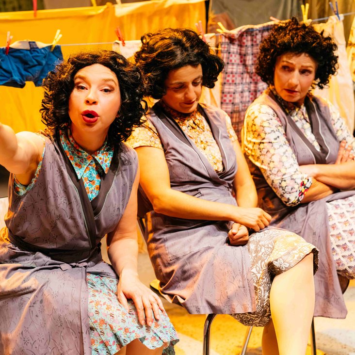 Drei Frauen sitzen nebeneinander auf der Bühne, die Frau im Vordergrund spricht gerade und zeigt mit ihrem rechten Arm auf etwas, Wäsche im Hintergrund