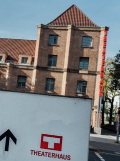 Der Blick auf das Theaterhaus auf dem Weg dorthin. Im Hintergrund sieht man das Theaterhaus. Im Vordergrund ist ein Wegweiser Schild, auf dem das Theaterhaus-Logo und ein Richtungspfeil zu sehen sind.