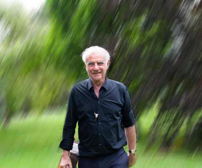 Roland Baisch in dunkler Kleidung läuft über eine grüne Wiese, im Hintergrund sind Bäume zu sehen. Er hat eine Gitarre in der Hand. Das ganze Bild ist etwas unscharf gestaltet.
