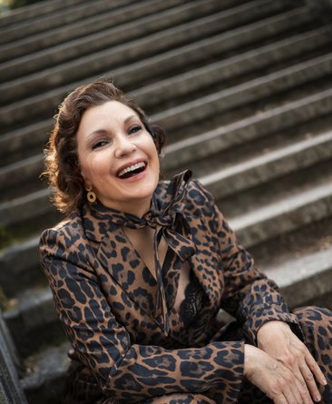 Portraitfoto Claudia Carbo, sie sitzt auf Treppenstufen und trägt Hose und Bluse im Leopardenmuster. Sie lacht in die Kamera.
