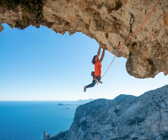 Alexander Huber hält sich mit beiden Händen an einem großen Felsvorsprung fest, im Hintergrund Felsküste und das Meer. Es ist mit einem Seil gesichert.