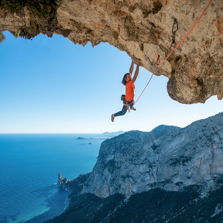Alexander Huber hält sich mit beiden Händen an einem großen Felsvorsprung fest, im Hintergrund Felsküste und das Meer. Es ist mit einem Seil gesichert.