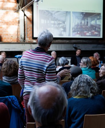 Bei der Mitgliederversammlung steht eine Person aus dem Publikum und spricht mit den Theaterhaus-Vertreter:innen auf dem Podium