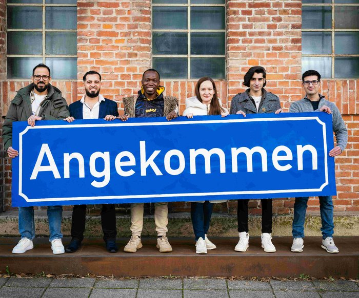 Die Teilnehmen der Produktion "Angekommen" stehen vor der Backsteinwand des Theaterhauses, sie halten alle gemeinsam ein großes blaues Schild mit der Aufschrift "Angekommen" in den Händen