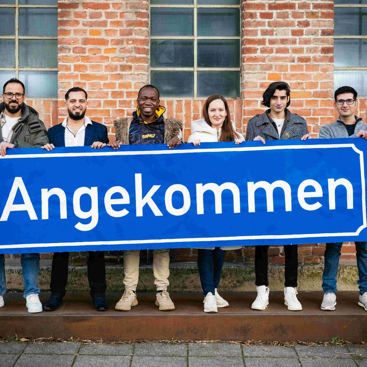 Die Teilnehmen der Produktion "Angekommen" stehen vor der Backsteinwand des Theaterhauses, sie halten alle gemeinsam ein großes blaues Schild mit der Aufschrift "Angekommen" in den Händen