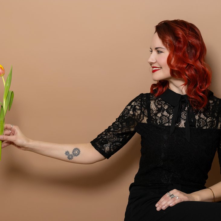 Portraitfoto von Sarah Straub. Sie hat rotes, lockiges Haar, trägt ein schwarzes Kleid und hat eine gelbe Blume in der Hand.