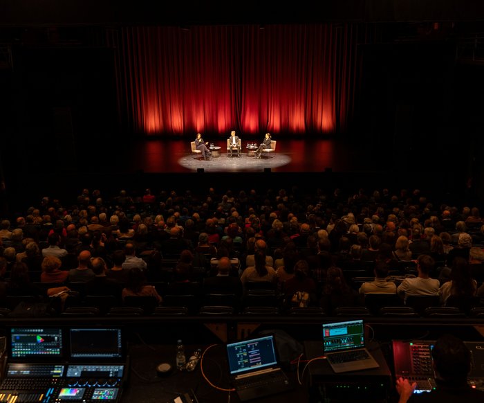 Ein Theatersaal, in den Rängen sitzen Menschen, im Hintergrund ist eine Gesprächssituation auf der Bühne. Im Vordergrund ist das Technikpult zu sehen.