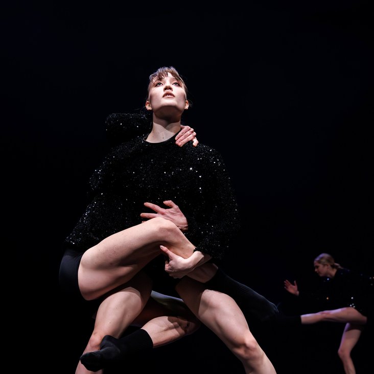 Tänzer:innen in Kostüm auf der Bühne, der Boden ist schwarz und die Bühne ist professionell ausgeleuchtet.