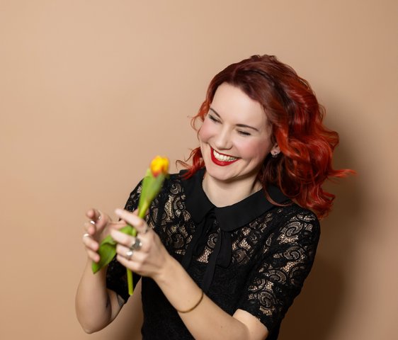 Portraitfoto von Sarah Straub. Sie hat rotes, lockiges Haar, trägt ein schwarzes Kleid und hat eine gelbe Blume in der Hand.