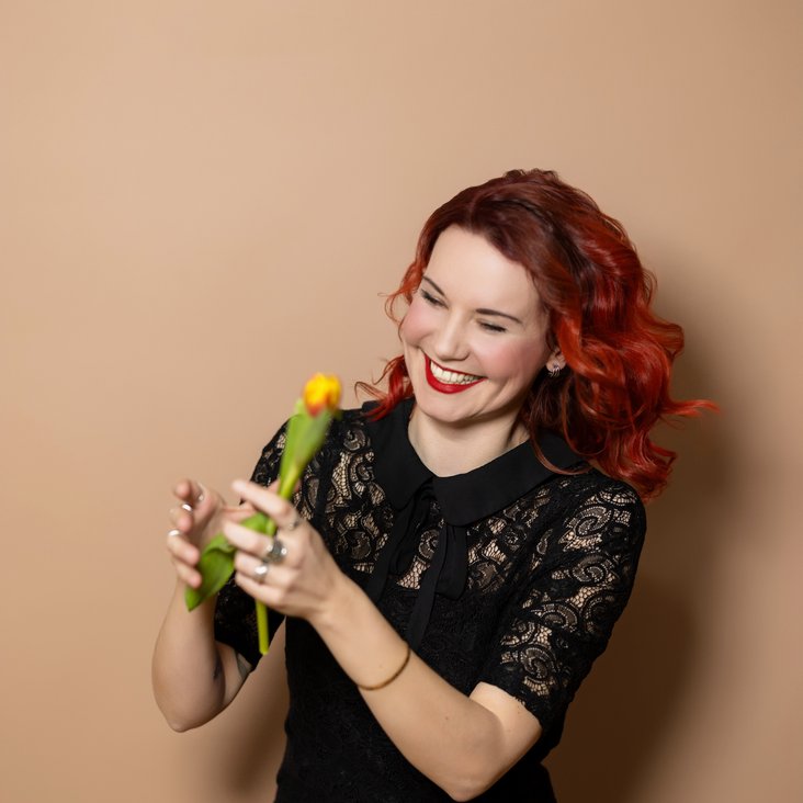 Portraitfoto von Sarah Straub. Sie hat rotes, lockiges Haar, trägt ein schwarzes Kleid und hat eine gelbe Blume in der Hand.