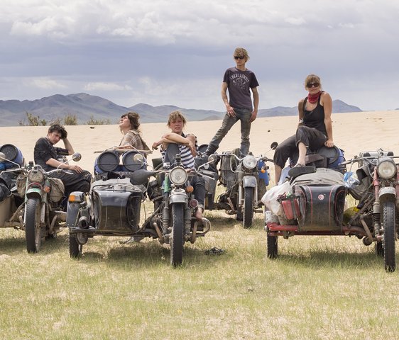 Fünf Personen stehen und sitzen auf ihren Motorrädern, die voller Gepäck sind auf einer Wiese mit Bergen im Hintergrund