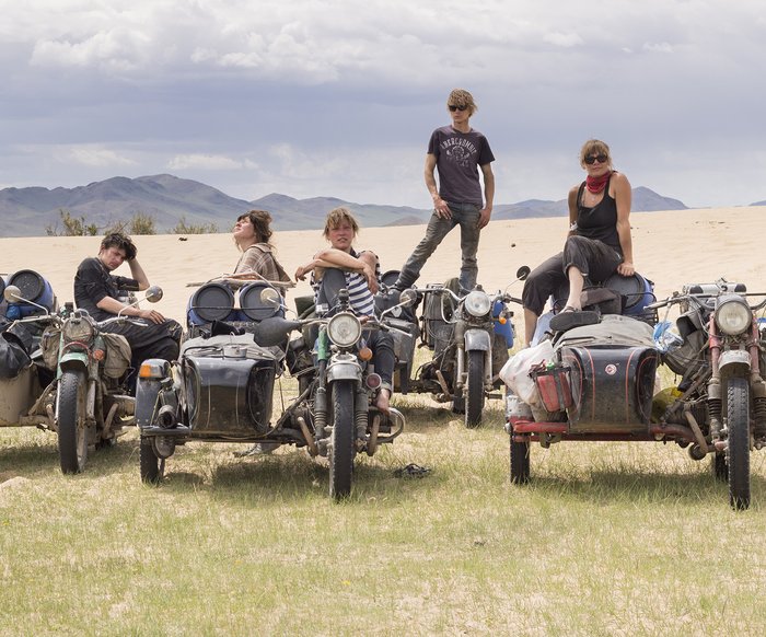 Fünf Personen stehen und sitzen auf ihren Motorrädern, die voller Gepäck sind auf einer Wiese mit Bergen im Hintergrund