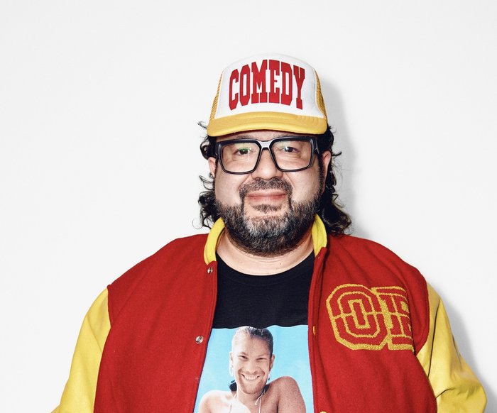 Der Künstler steht in einer geld-roten Collagejacke und einer Cap da und lächelt in die Kamera.