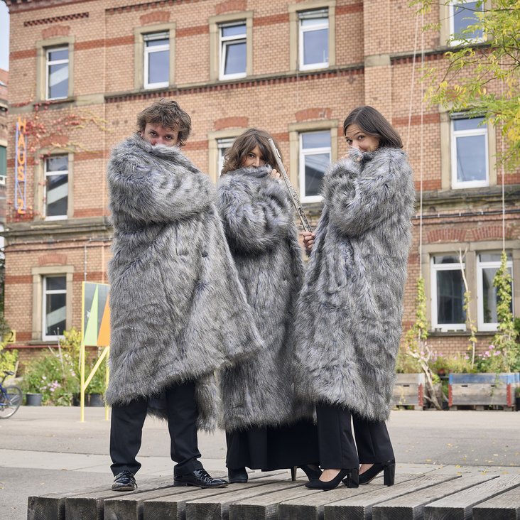 Drei Menschen stehen vor einem Backsteingebäude. Sie tragen schwarze Hosen und lange, graue Mäntel in Felloptik.