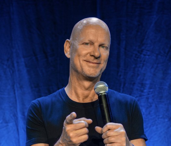 Rüdiger Hoffmann live auf der Bühne. Er trägt ein schwarzes T-Shirt und hat ein Mikrofon in der Hand. Er zeigt mit dem Zeigefinger in Richtung der Kamera und grinst.