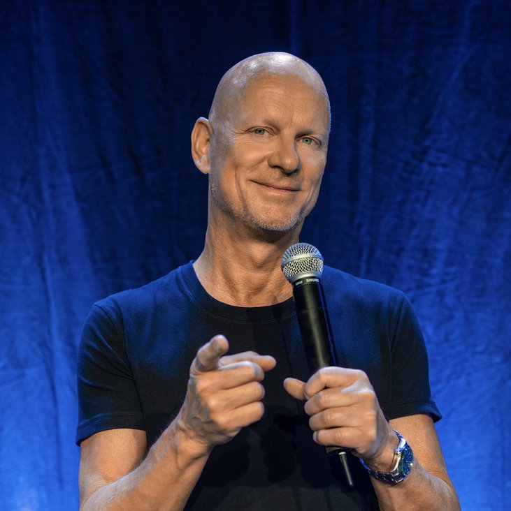 Rüdiger Hoffmann live auf der Bühne. Er trägt ein schwarzes T-Shirt und hat ein Mikrofon in der Hand. Er zeigt mit dem Zeigefinger in Richtung der Kamera und grinst.