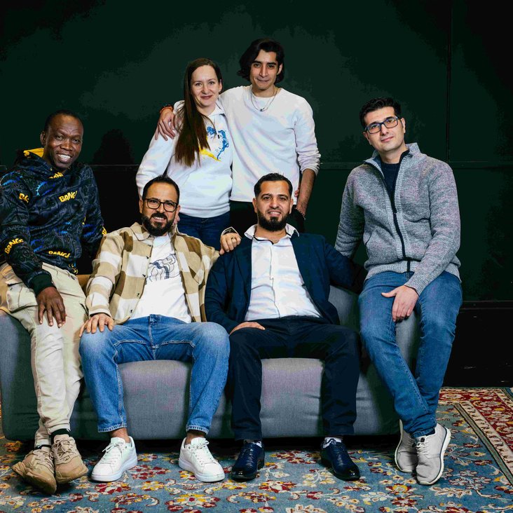 Die Teilnehmenden der Produktion "Angekommen" stehen hinter und sitzen auf einem Sofa, das auf einem bunten Teppich steht.