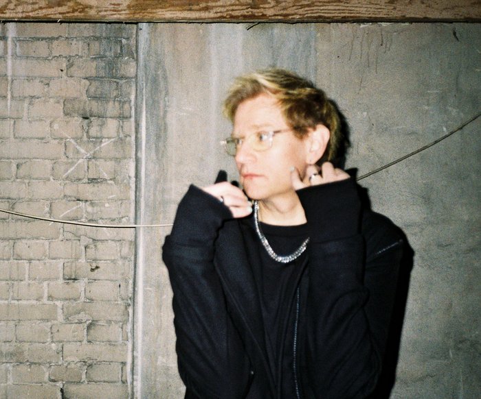 Porträt einer Person in dunkler Kleidung vor Backsteinwand, Hände am Kragen, seitlicher Blick.