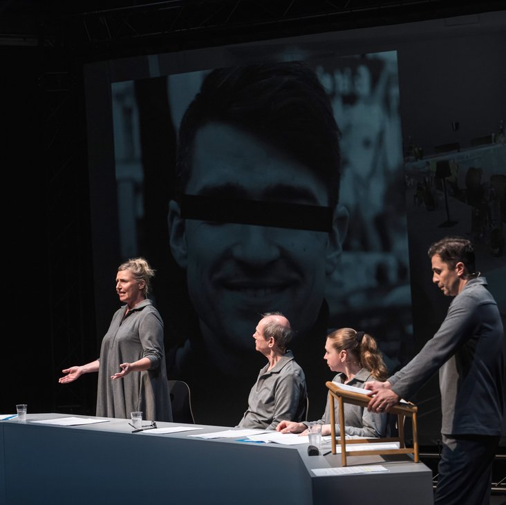 Szenenfoto, es zeigt fünf Schauspieler:innen sitzend und stehend in grauer Kleidung auf der Bühne. Im Hintergrund das schwarz-weiß Foto einer Person, die Augen sind zensiert.