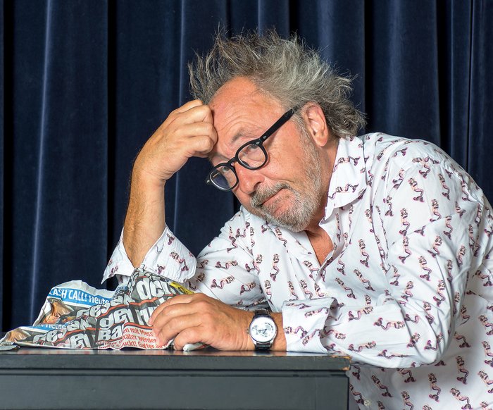 Portraitfoto Urban Priol. Er sitzt, den Kopf aufgestützt, mit einer Tageszeitung an einem Tisch auf der Bühne.